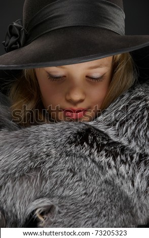 Portrait of a beautiful young lady in grey fur coat and black hat looking down on a dark background