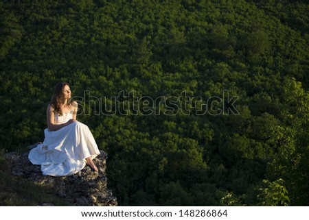 Beautiful young woman wearing elegant white dress standing with a smile on a road in the forest with rays of sunlight beaming through the leaves of the trees
