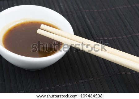 Hoisin & Chopsticks - Close up of chopsticks resting on a small white bowl of asian dipping sauce on a black mat.