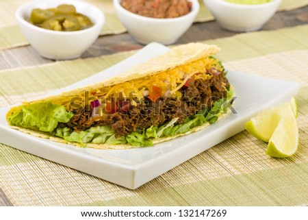 Beef Tacos - Mexican shredded beef tacos in soft corn tortillas served with lettuce, sour cream, grated cheddar cheese and salsa. Guacamole, refried beans and jalapeÃ?Â±os on the background.