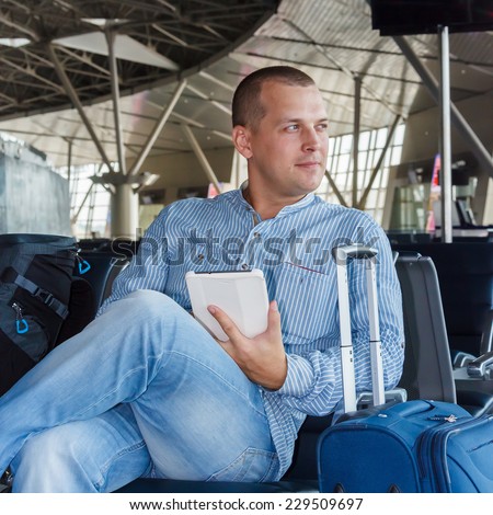handsome young man with short hair working on the tablet, sitting on a chair with things at the airport waiting for his flight