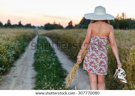 Portrait of young pretty girl at summer weather dressed in bright hat goes on road through a field of wheat