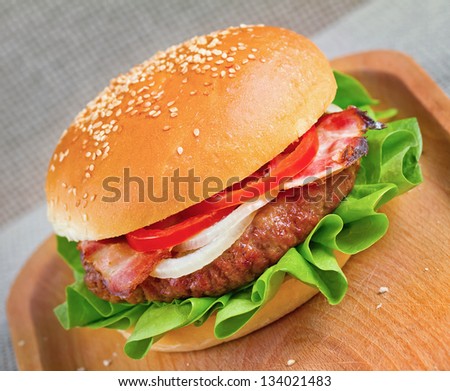 The hamburger, a ground beef patty between two slices of bread with lettuce, onion, bacon and tomato on wooden plate