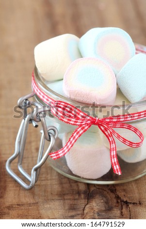 Pastel colour marshmallows in a glass jar decorated with a red checkered bow against rustic wooden background