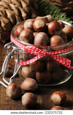Shelled hazelnuts in a glass jar decorated with a red Christmas ribbon on a rustic wooden kitchen table with pine cones at the background