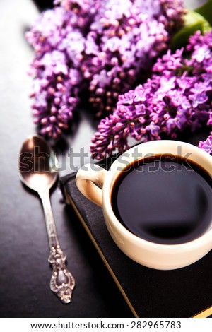 Black coffee in a cup, book, a spoon and fresh lilac flowers on black background