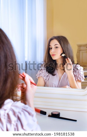 Portrait of  beautiful young woman looking at the mirror putting on makeup holding a makeup brush