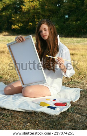 Young lady with long brown hair and wearing a white shirt seen smiling as she paints on a canvas. with a curious look