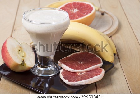 Smoothie of banana, apple and grapefruit with yogurt in a cup on a brown plate and fruit on the table next.