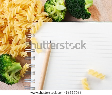 Directly above close-up view of a table with a pasta, broccoli and a notebook on cutting board