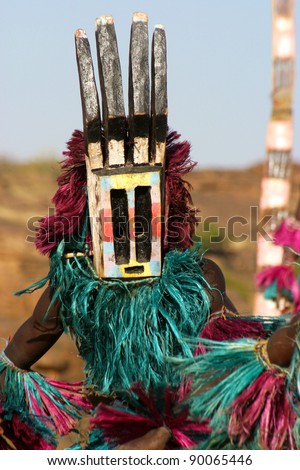 Dogon dancer with mask, representing a deer (antlers), with on the background a so-called tall mask which can be several meters high