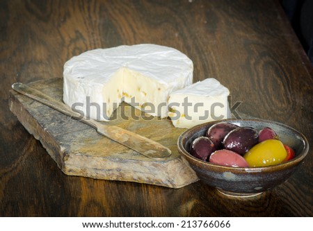 Brie cheese with a wedge cut out on a slate platter with a tarnished vintage knife and olives in a pottery bowl