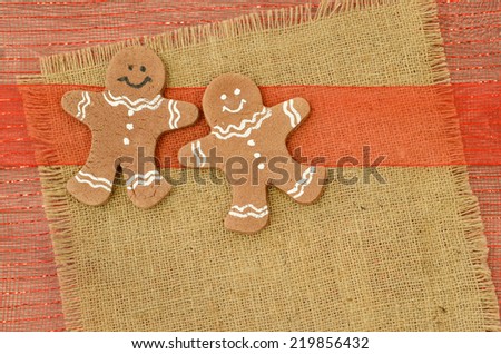 Two gingerbread men on a square of burlap with a red sheer ribbon and a Christmas mesh background