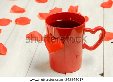 A red mug with a heart shaped handle with a heart attached to a tea bag on a rose petal strewn surface