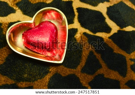 A foil wrapped red chocolate in a heart shaped brass dish on a faux animal skin surface