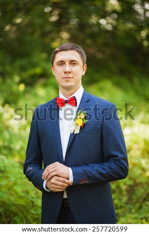 portrait of handsome young man smartly dressed in blue suit, white shirt and red bowtie. groom ready for wedding celebration standing over green nature background and waiting for bride