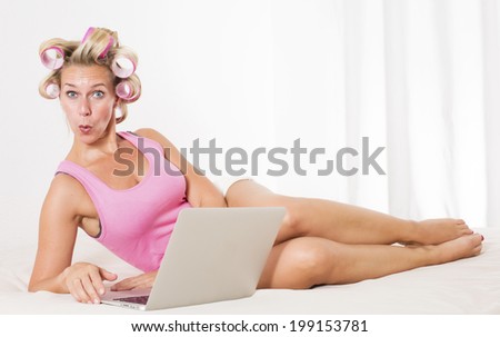 blond woman with pink curlers laying in bed with a laptop