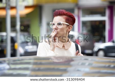 portrait of a red-haired woman standing behind a car and smoking a cigarette