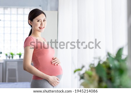 Side view of beautiful asian pregnant woman standing near window and smile at you in the living room Imagine de stoc © 