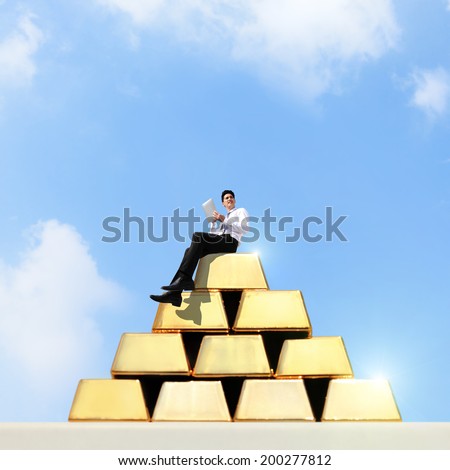 Successful business man working and using tablet pc on stacks and rows of gold ingots with sky