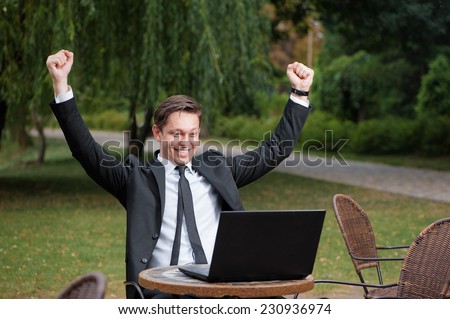 Business winner. Happy young caucasian man in formal wear keeping arms raised and expressing positivity while sitting a the  outdoors cafe working with laptop with park in the background