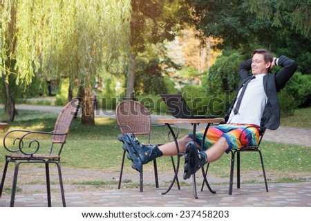 Work and relax outdoor. Businessman dressed in suit, shorts and rollers working with laptop at the park cafe
