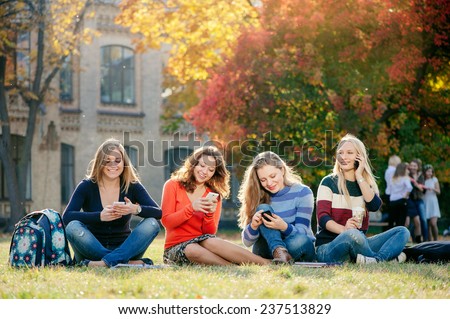 Friendship, leisure, technology and people concept - group of smiling girl friends with smartphones sitting on grass with university building in the background