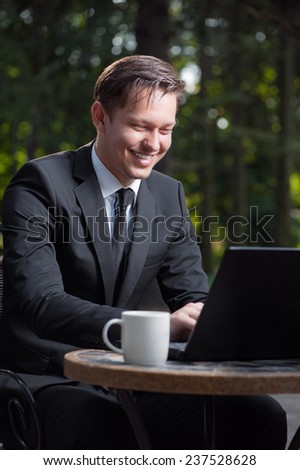 Successful businessman. Attractive young caucasian man in formal wear working on laptop and smiling while sitting at the table outdoors.