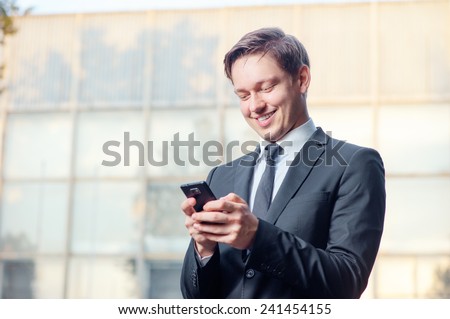 Typing business message. Cheerful young man in formal wear holding mobile phone and smiling while standing outdoors