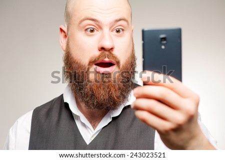 It is unbelievable! Surprised young man in shirt looking at mobile phone and keeping mouth open while standing against grey studio background