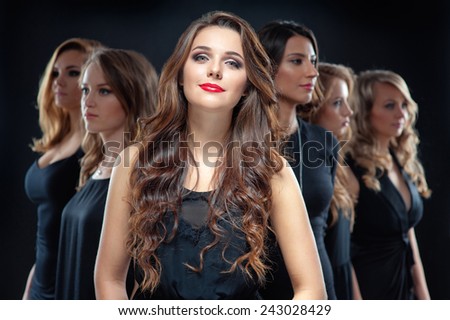 Group of beautiful women. Attractive young woman in black dress with long hair looking at camera and her friends standing in front her back against black background.