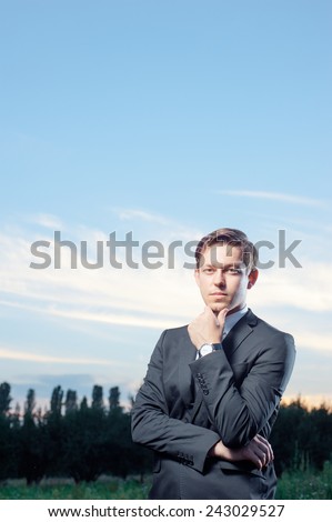 In search of the new ideas. Thoughtful young caucasian man in formal wear holding hand on chin and looking at camera against sky background.