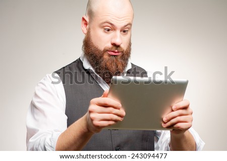Shocking news online. Surprised young caucasian man with long beard holding digital tablet while standing against studio background