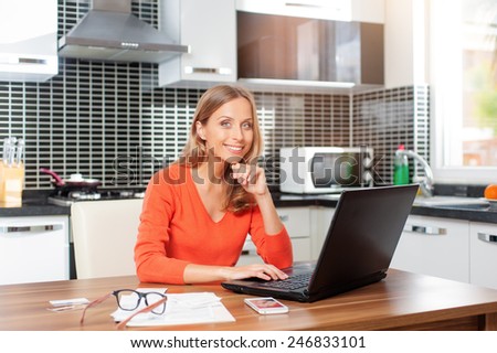 Lovely blonde woman working with her laptop while sitting in the kitchen in her apartment holding her chin and looking at camera