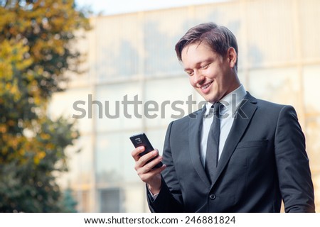 Typing business message. Cheerful young man in formal wear holding mobile phone and smiling while standing outdoors