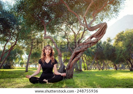 Yoga outdoors. Beautiful young caucasian woman doing lotus position yoga in park on green grass under the tree.
