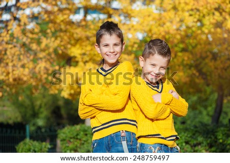 Two smiling brothers in the park on an autumn day standing back to back with crossed arms and looking at camera.