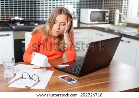stressed over bills. portrait of a young caucasian woman using a laptop computer sitting at her kitchen holding utility bills and bank statements being thoughtful and worried. Home interior.