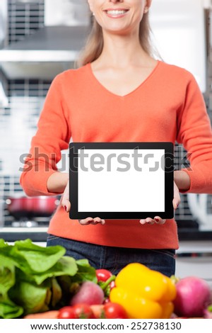 Copy space at her tablet. Cheerful young woman holding digital tablet standing in her kitchen at home
