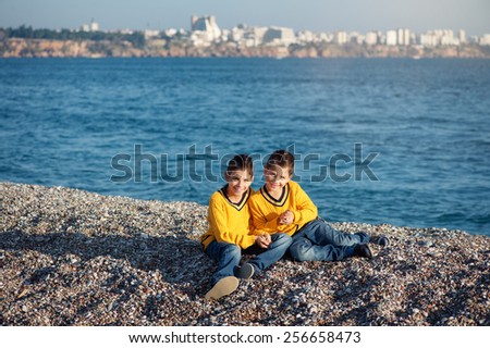 A portrait of two happy, smiling young brothers, a little child and his toddler brother, sitting outside on the pebble beach against the sea