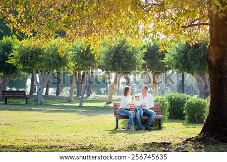 Smiling family in autumn park. Happy young parents with little kid sitting on the bench under the tree.