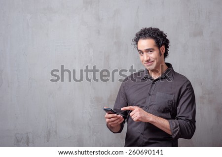 Typing message. Handsome young man in shirt holding mobile phone and looking at camera while standing against grey background