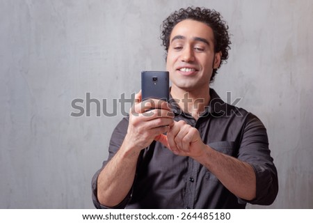 Handsome young man in shirt holding mobile phone and taking photo by phone camera while standing against grey background