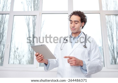 Doctor with digital tablet. Confident young Arabic man in medical uniform working on digital tablet while standing against window.
