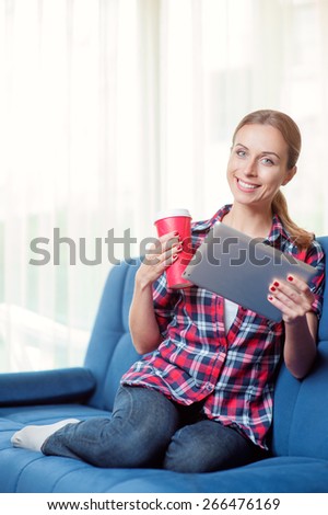 Working at home. Beautiful young smiling woman using tablet pc while drinking coffee sitting on sofa at home looking at camera.