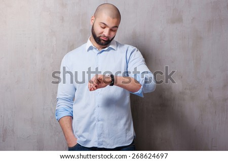 It\'s time! Yong handsome bearded man looking at his hand watch while standing against grey wall.