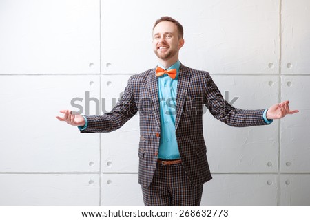 You are welcome! Cheerful young man in plaid suit and bowtie gesturing welcome sign and smiling while standing against white wall.