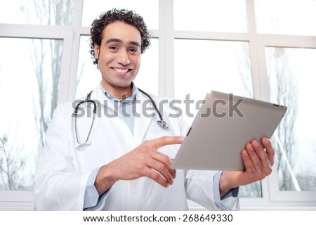 Doctor with digital tablet. Smiling young Arabic man in medical uniform working on digital tablet while standing against window.