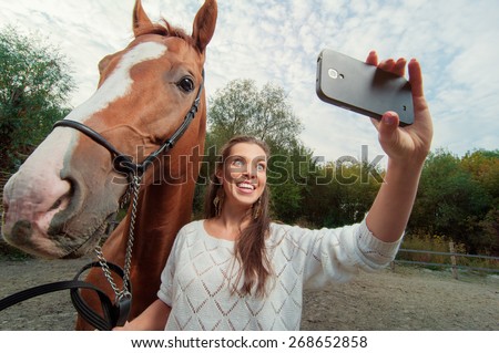 Funny selfie with my friend! Attractive smiling young woman holding smartphone and making selfie with her horse outdoors