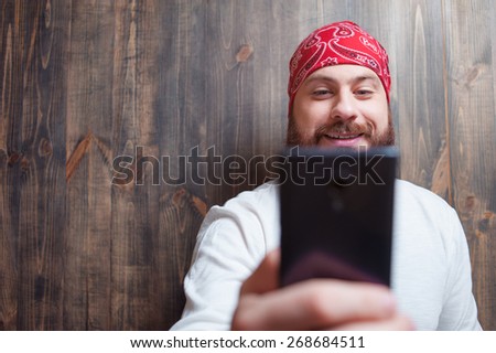 Young smiling bearded man  holding mobile phone and looking at it against wooden wall.